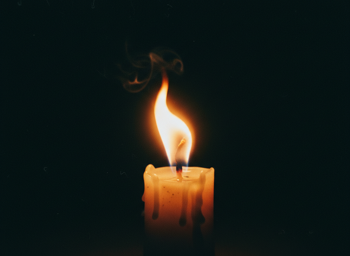 Moody close-up photograph of a single flickering candle flame in near-total darkness, warm orange glow bleeding into deep black shadows.